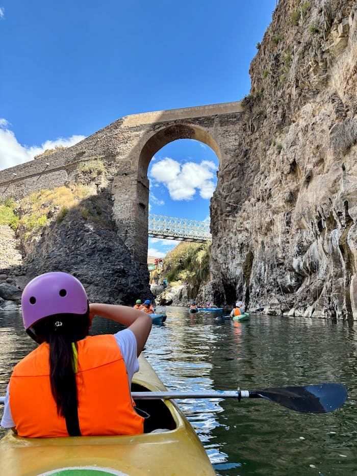 A group kayaking under a historical stone bridge in a scenic gorge with clear blue skies.